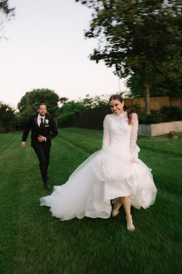 Novios caminando juntos por el césped de un jardín, ella recogiéndose la falda del vestido
