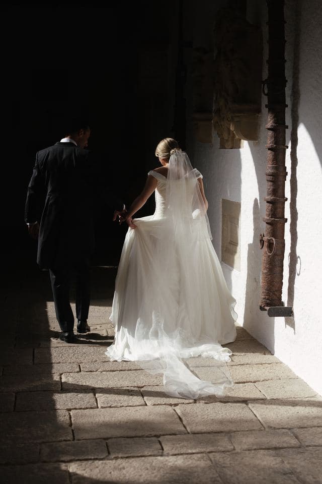 Los novios caminan de espaldas por un claustro de piedra con la luz entrando de lado