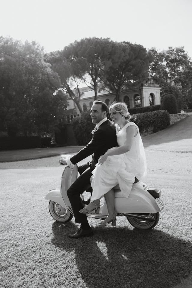 Los novios ríen montados en una vespa clásica en el jardín de una finca, en blanco y negro