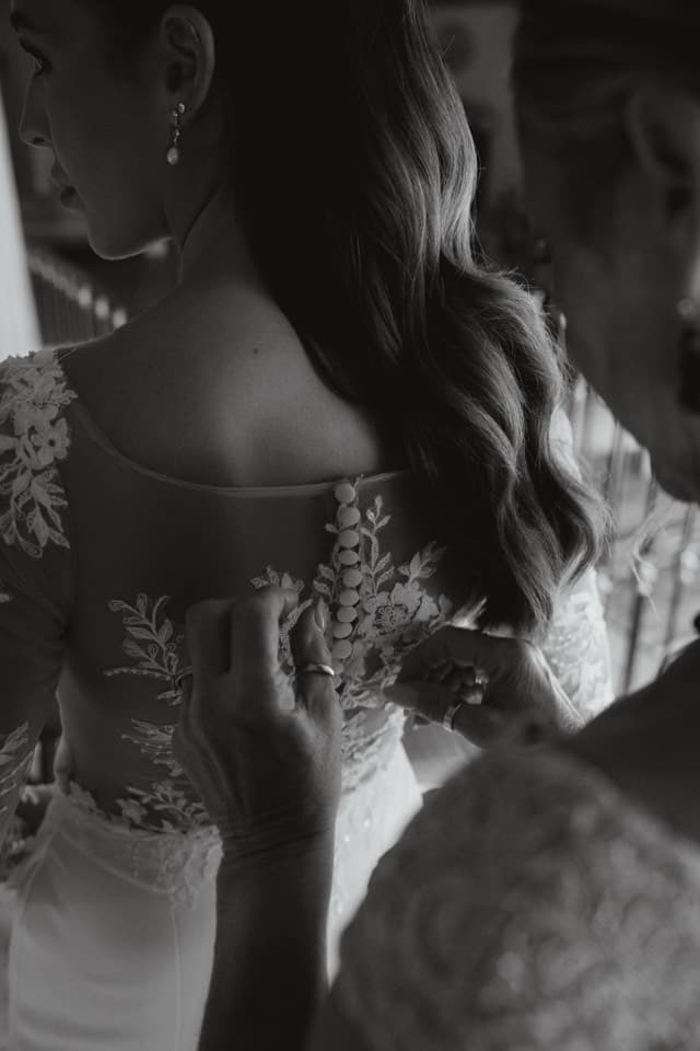 Unas manos abotonan la espalda de encaje del vestido de la novia, en blanco y negro